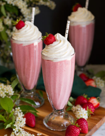 Three homemade strawberry milkshakes in tall glasses topped with whipped cream and strawberries.
